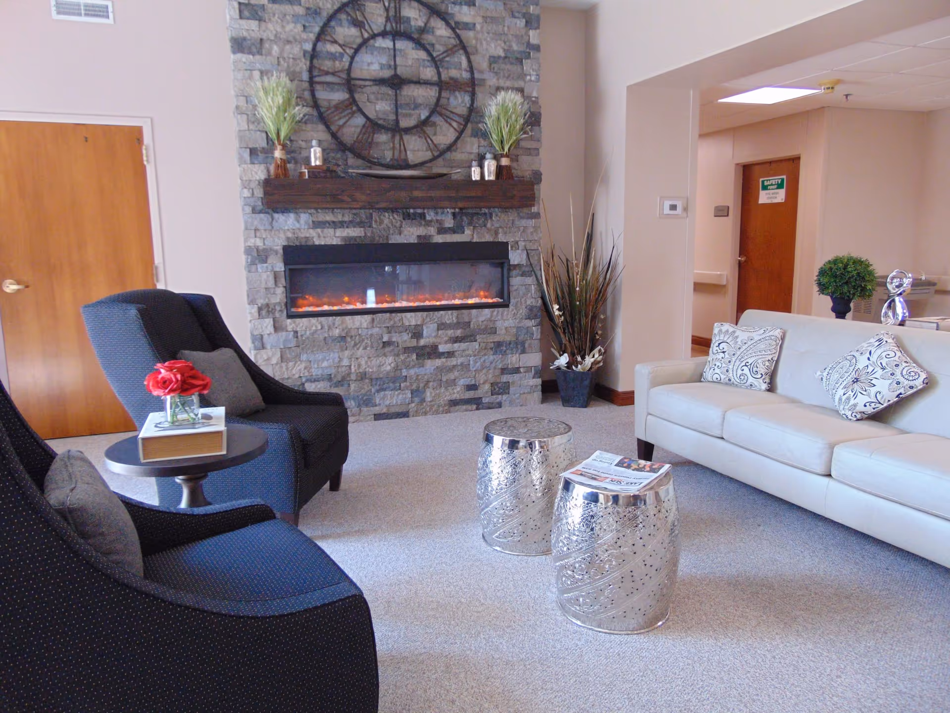 A cozy living room area in Good Shepherd Care Center featuring a stone fireplace with a large decorative clock above it. There are two dark blue armchairs with gray pillows, a small round side table with a book and a vase of red flowers, two silver decorative stools with a newspaper on one, and a light-colored sofa with patterned cushions. The room has beige walls, carpeted floor, and a hallway with a wooden door labeled 'Safety First'.