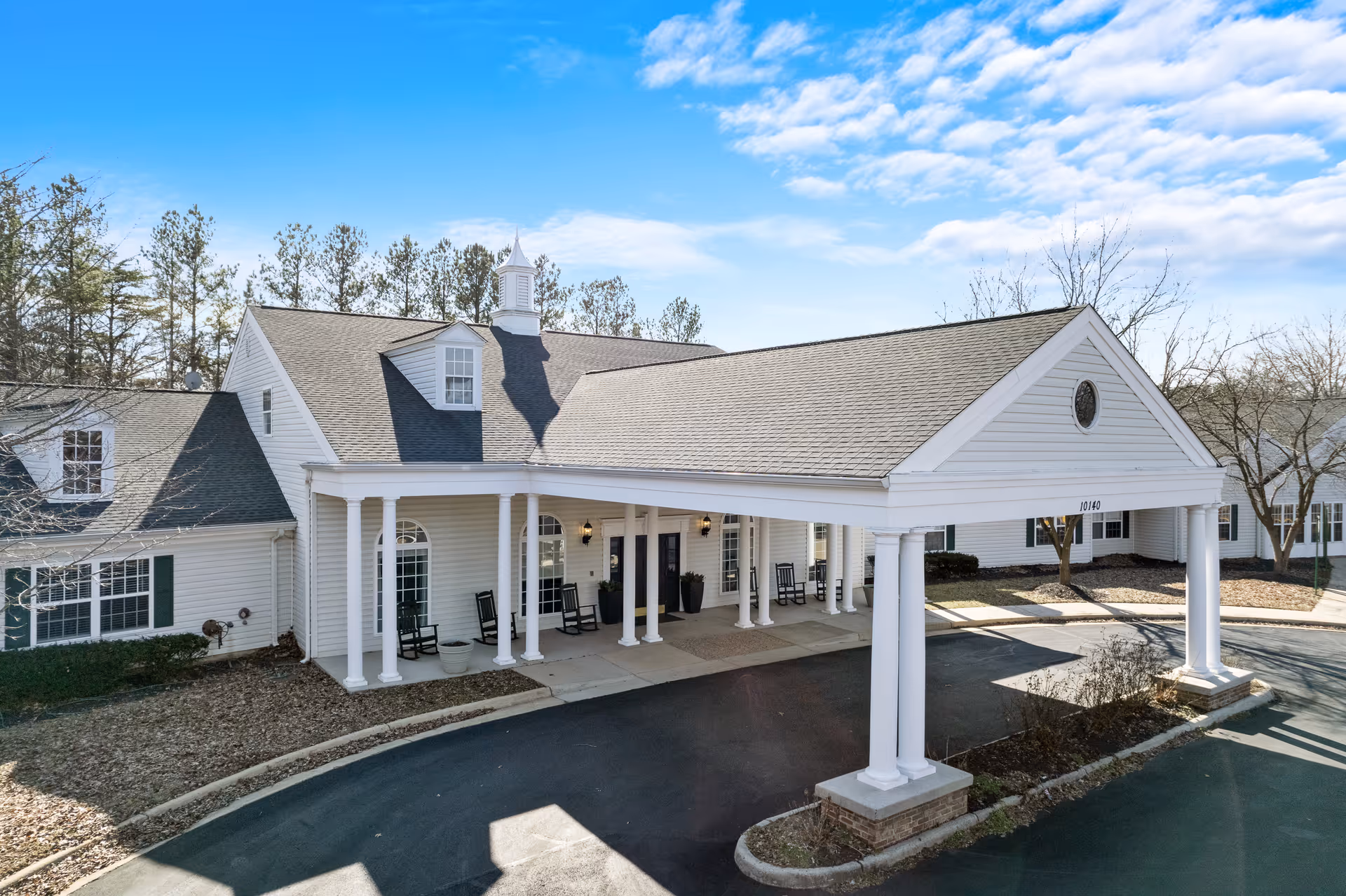 Front entrance of a white, colonial-style building with a covered porte-cochere supported by columns and rocking chairs on the porch.