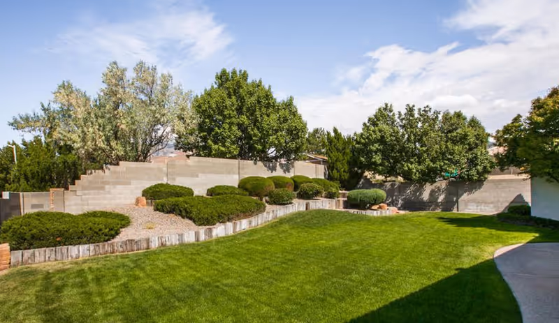 A well-maintained outdoor garden area with green grass, trimmed bushes, and several trees. There is a concrete block wall in the background under a partly cloudy blue sky.
