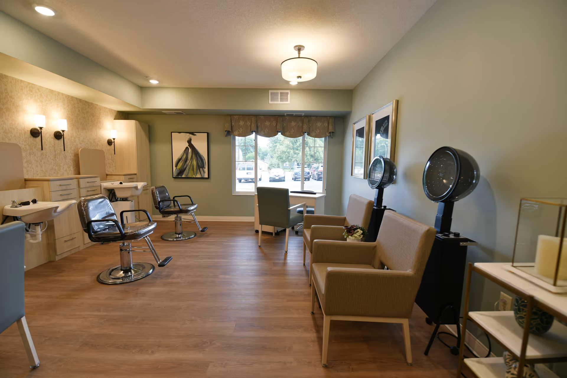 Interior view of a hair salon area in a senior living facility with two salon chairs and sinks on the left, two hair dryers on the right, beige armchairs, a desk with a chair near a window, and framed artwork on the walls.