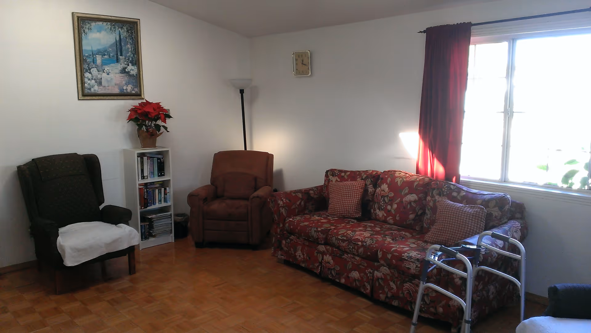 A cozy living room with a floral patterned sofa, two armchairs, a small white bookshelf with books and a potted poinsettia plant on top, a floor lamp, a wall clock, a large window with red curtains, and a walker placed near the sofa.