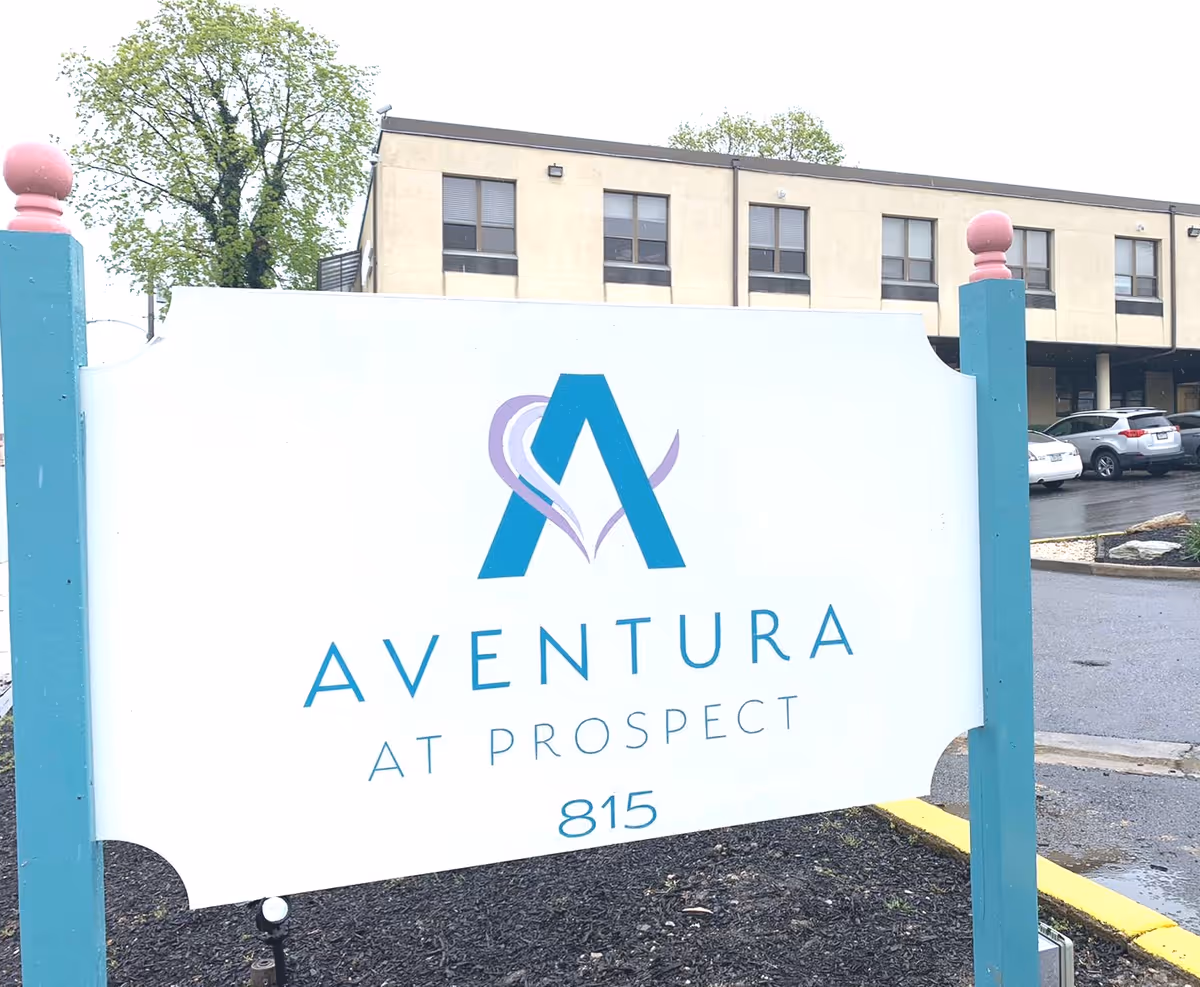 Outdoor image showing a white sign with teal posts and pink finials for Aventura At Prospect, with a building and parked cars in the background on a cloudy day.