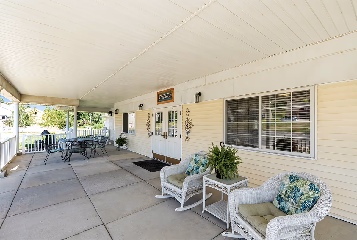 Covered front porch with wicker chairs, a small table and an outdoor dining set outside the entrance of a senior living building.