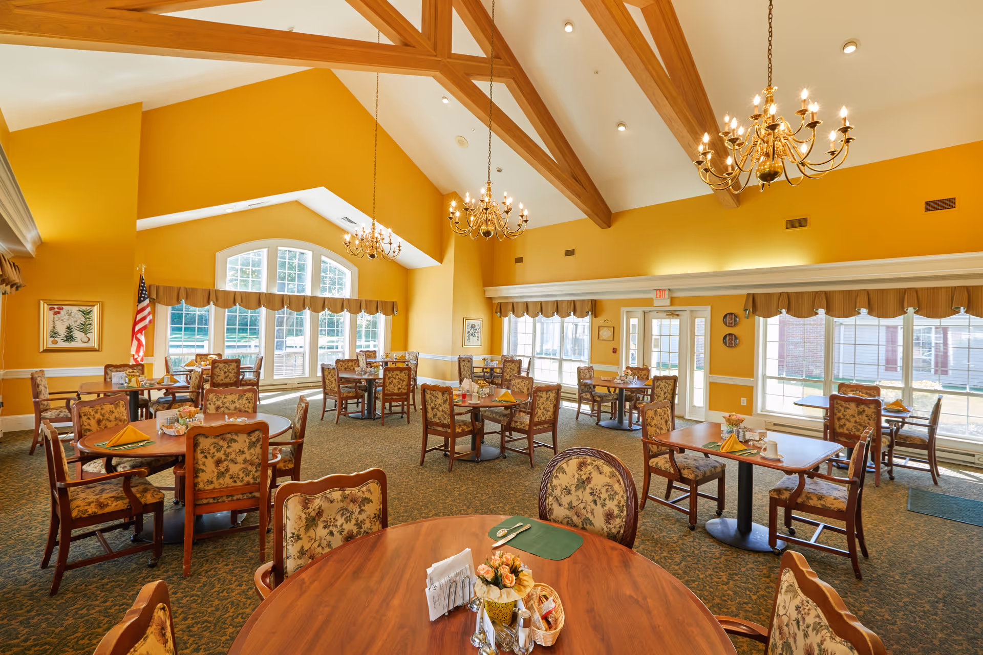A spacious dining room with yellow walls and large windows letting in natural light. The room features multiple round and square wooden tables with floral upholstered chairs. Chandeliers hang from the ceiling with exposed wooden beams. The tables are set with placemats, napkins, and small flower arrangements. An American flag is visible near the windows.