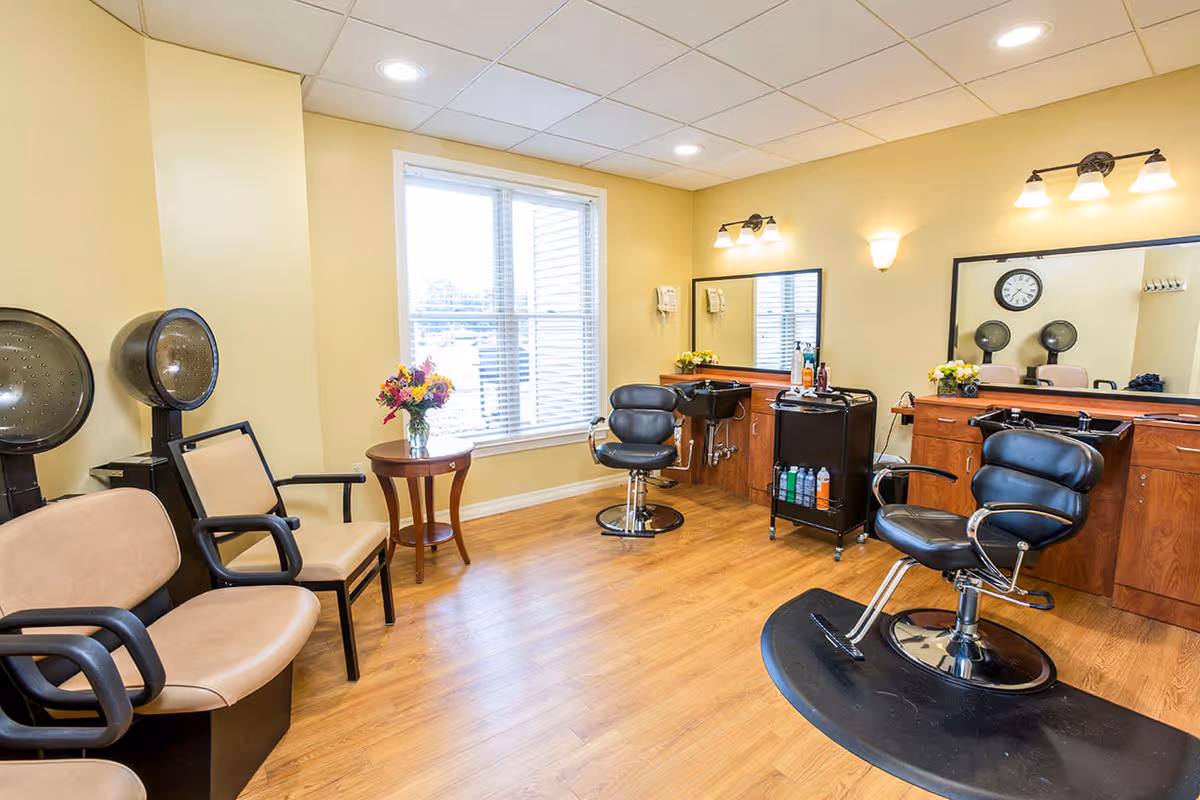 A bright hair salon room with wooden flooring and beige walls. The room features two black salon chairs in front of large mirrors mounted on wooden cabinets. There are two hair dryers with chairs on the left side, a small round table with a vase of colorful flowers near a window with white blinds, and a rolling cart with hair products between the salon chairs. The room is well-lit with ceiling lights and wall sconces.