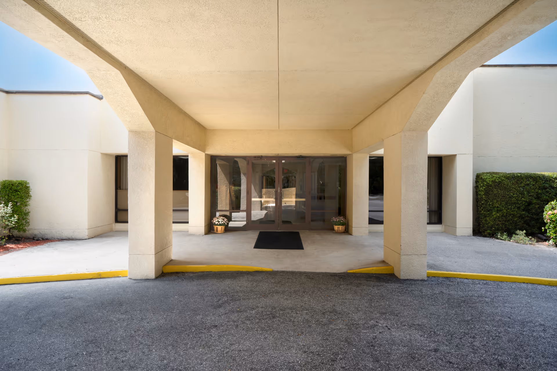Covered entrance area of a building with beige walls and pillars, a black doormat in front of glass double doors, and two potted plants with flowers on either side of the entrance.