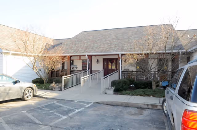 Front entrance of a single-story building with a sloped roof, featuring a wheelchair accessible ramp and handrails. There are leafless trees and shrubs on either side of the entrance, and two parked cars are visible in the parking area in front of the building.