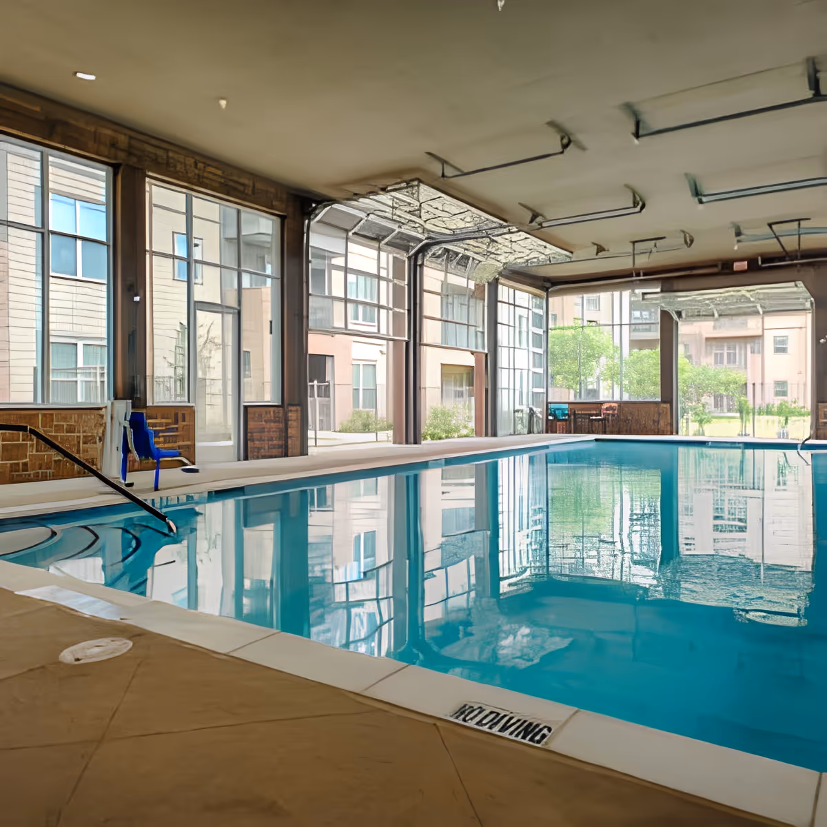 Indoor swimming pool area with clear blue water, large windows letting in natural light, and a blue pool lift chair on the side. The pool is surrounded by a tiled floor and the ceiling has exposed beams and lighting fixtures.