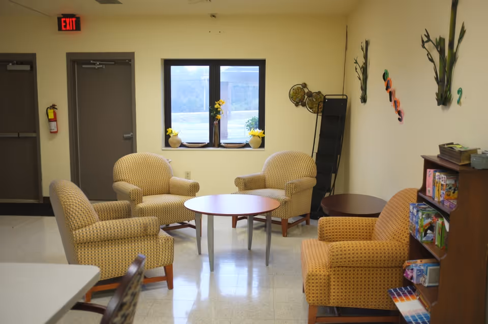 A cozy sitting area in an assisted living facility with four patterned armchairs arranged around a round wooden table. There is a window with three vases of yellow flowers on the windowsill. The walls are decorated with some plants and colorful letters. A bookshelf with board games and puzzles is visible on the right side. Two closed doors and an exit sign are visible in the background.