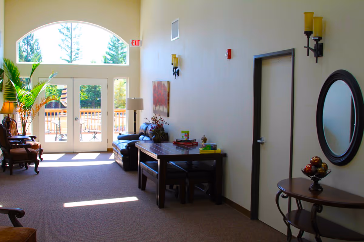 A bright and spacious senior living common area with large arched windows and glass doors letting in natural light. The room features a leather couch, a wooden table with benches, two armchairs, a floor lamp, a round mirror on the wall, and decorative plants. Outside the glass doors, a patio with chairs is visible.