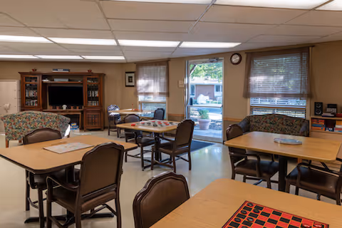 A common room in a senior living facility with several tables and chairs arranged for games and social activities. There is a large wooden entertainment center with a television, a floral-patterned loveseat, and windows with blinds letting in natural light. A door leads outside to a patio area.