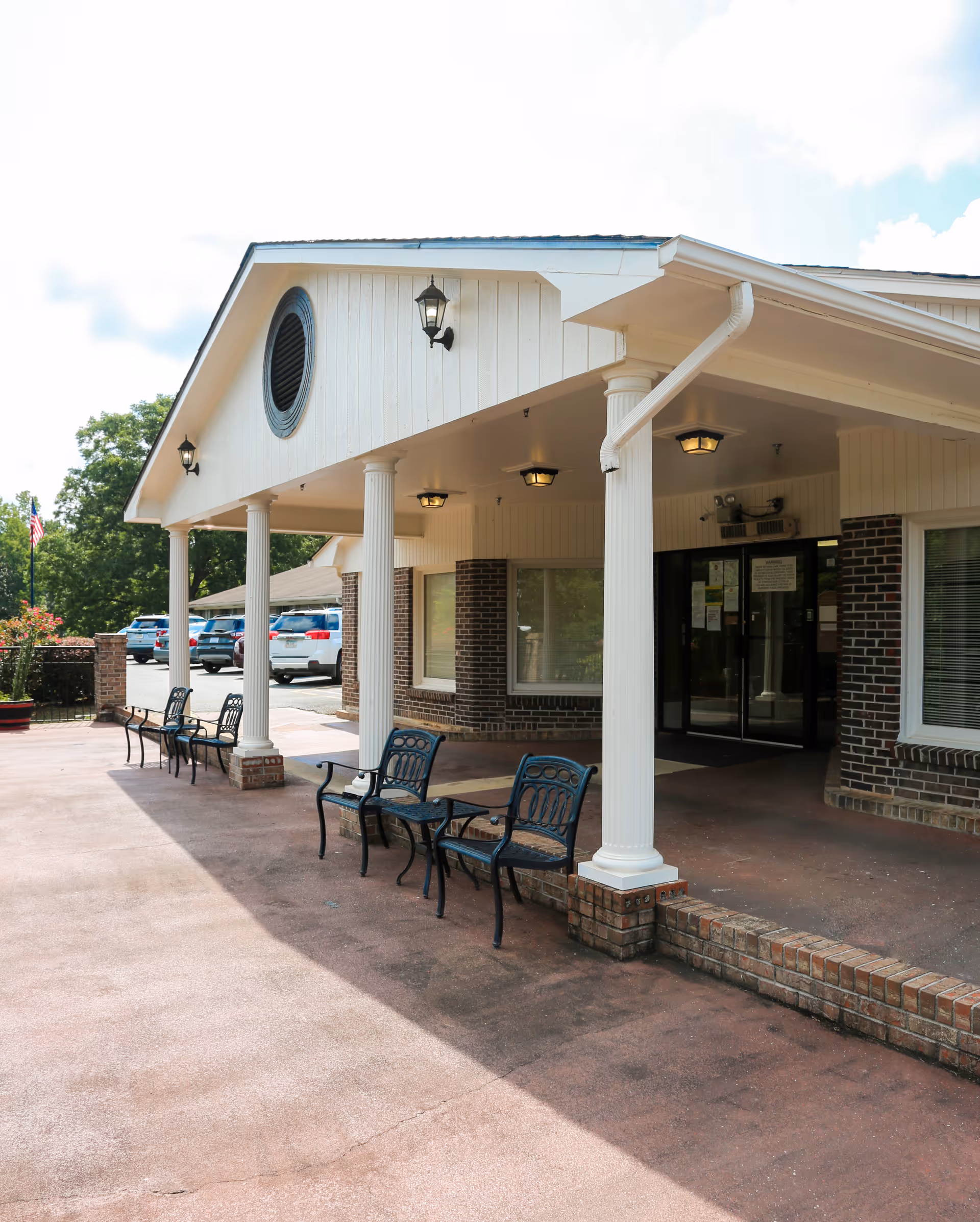 Covered front entrance of a brick senior living building with white columns, outdoor benches, and parked cars.