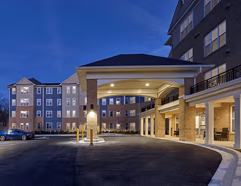Exterior view of a multi-story senior living facility at dusk with a covered entrance and well-lit windows. The building features brick and siding, with a driveway and parked cars visible.