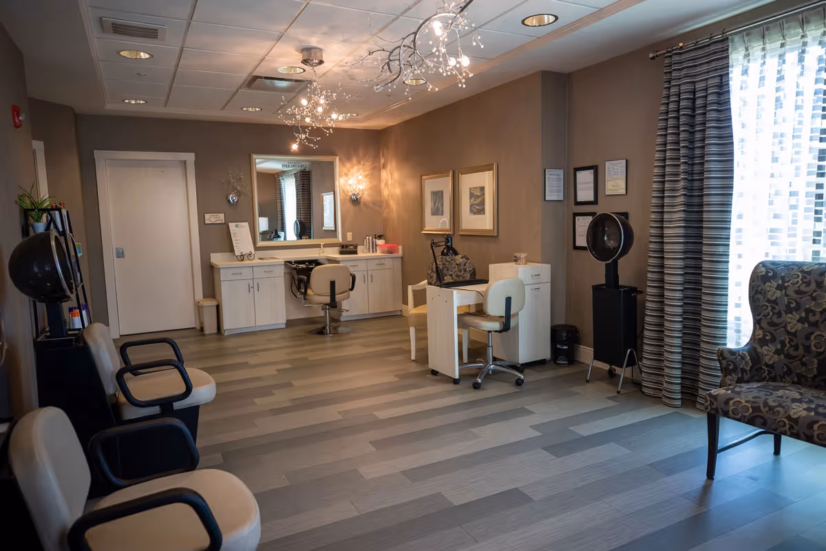 Interior view of a salon area in a senior living facility with beige walls, modern light fixtures, a large mirror above a counter with cabinets, salon chairs, a small desk with chairs, a patterned armchair near a window with striped curtains, and wood-look flooring.