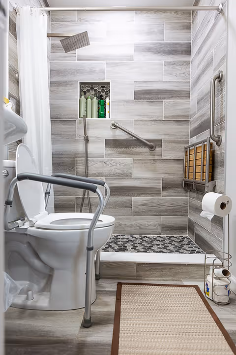 Accessible bathroom with a toilet equipped with support rails, a walk-in shower with a handheld showerhead, grab bars on the wall, and a small bench. The walls and floor are tiled in shades of gray and beige, and there is a white shower curtain. A toilet paper holder is mounted on the wall, and a beige rug with a brown border is on the floor.