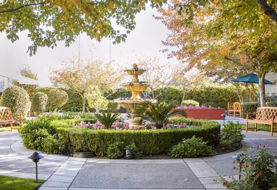 A peaceful outdoor garden area with a three-tiered stone fountain at the center surrounded by neatly trimmed hedges and colorful flowers. Wooden benches are placed along the paved pathways, and trees with green and autumn-colored leaves provide shade. The setting appears calm and inviting.