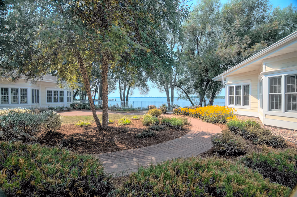 Pathway curves between pale buildings, with trees, shrubs, and a lake view beyond.