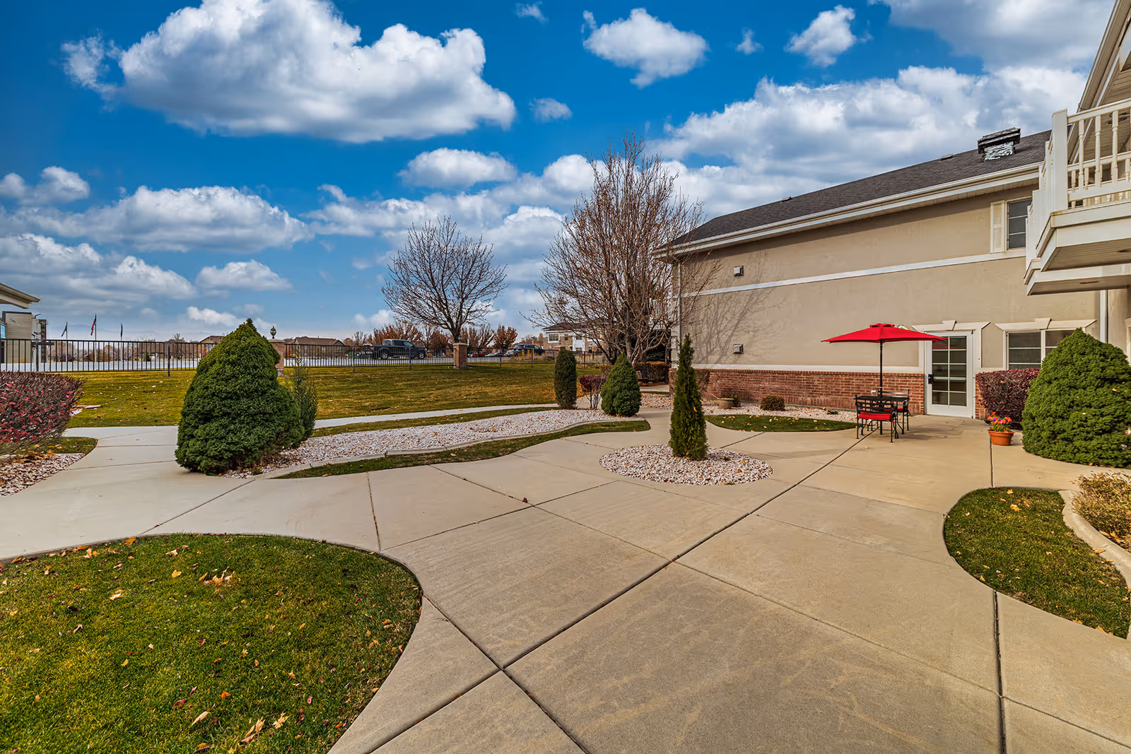 Exterior courtyard with paved walkways, landscaped shrubs, and a patio table with a red umbrella beside a building.