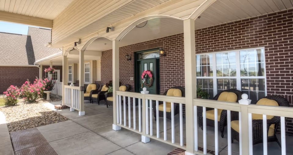 Covered porch area with a white railing and several wicker chairs with yellow cushions. The porch is attached to a brick building with a green door decorated with a pink floral wreath. Pink flowering bushes and a birdbath are visible near the porch.