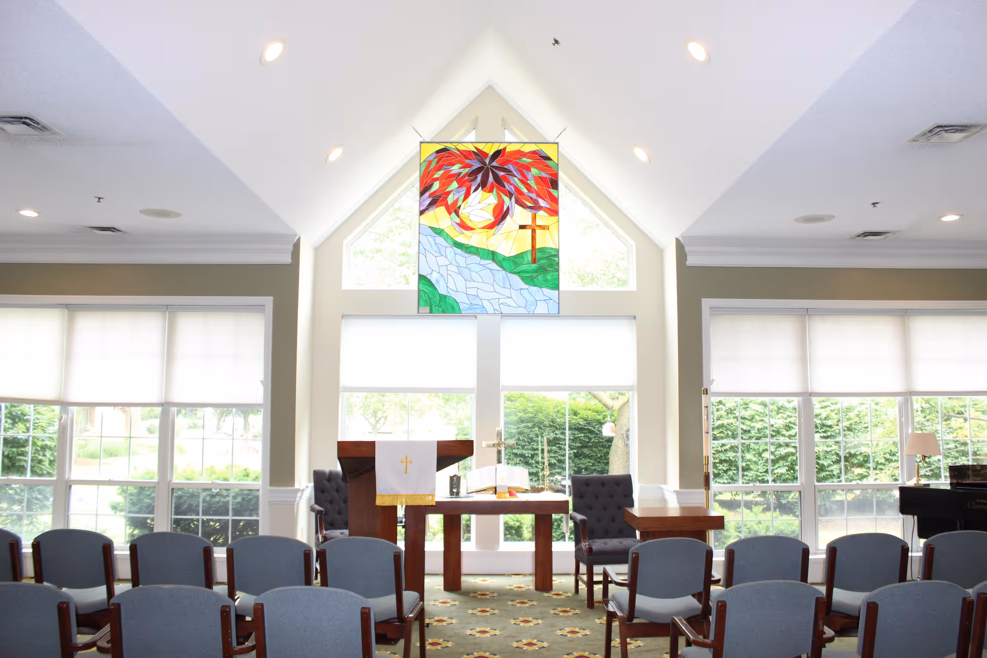 Interior view of a chapel or worship room with rows of chairs facing a wooden altar and lectern. A colorful stained glass window depicting a cross and a crown of thorns is above the altar. Large windows with blinds allow natural light to fill the room.