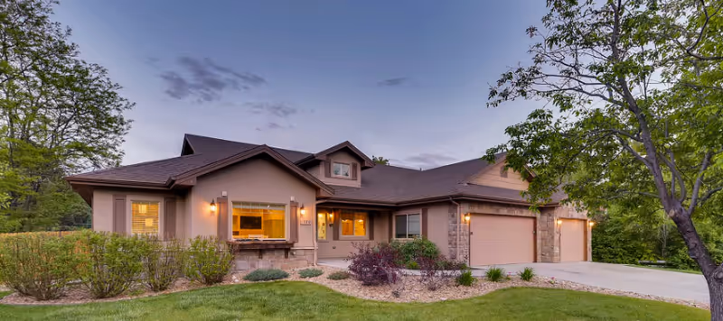 Exterior view of a single-story residential building with a three-car garage, stone and beige siding, surrounded by green trees and shrubs under a twilight sky.