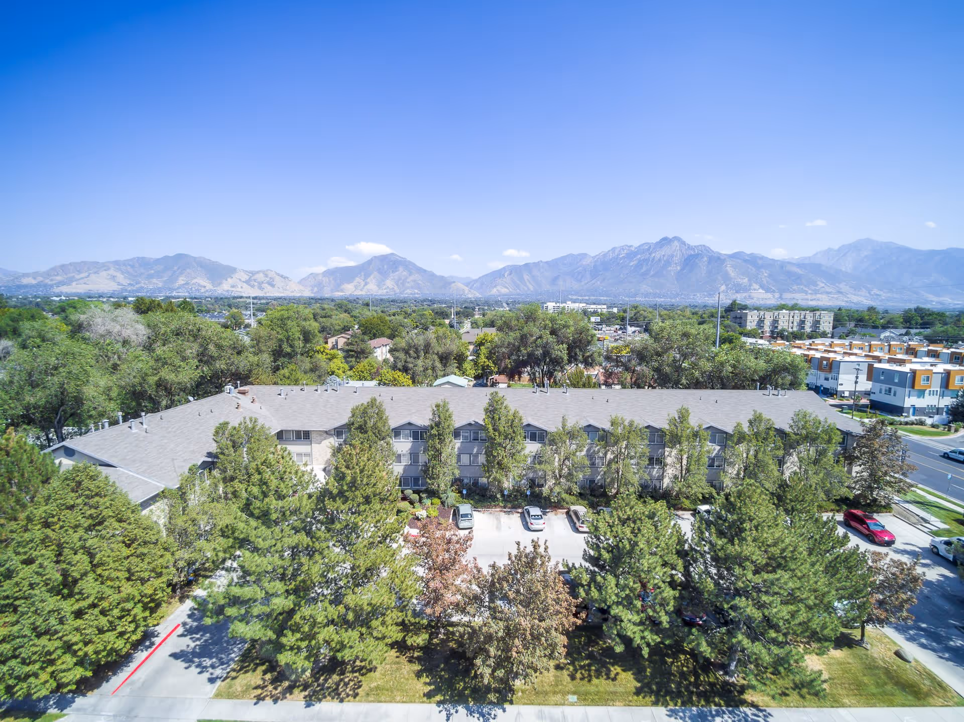 Aerial view of The Laurel at Millcreek senior living facility surrounded by trees with a parking lot in front and mountains in the background under a clear blue sky.