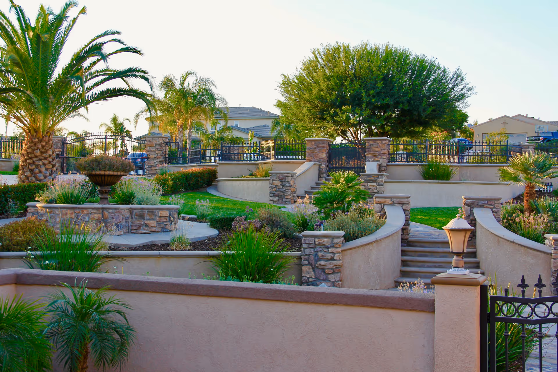 Outdoor landscaped garden area with stone walls, stairs, palm trees, and various plants under a clear sky at Pacific Vista Senior Living.
