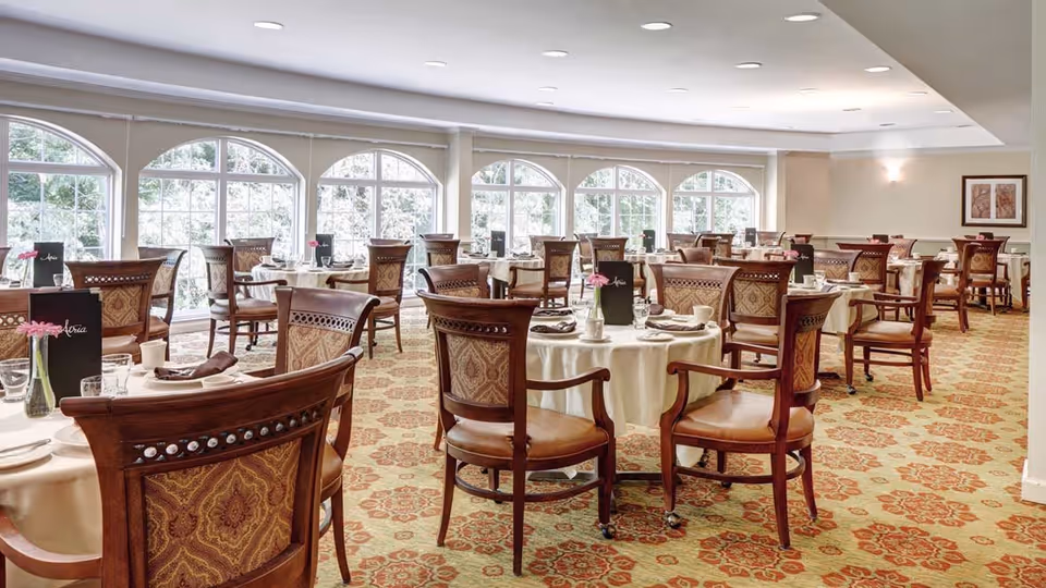 A spacious dining room with multiple round tables covered with white tablecloths, each set with plates, cups, napkins, and menus. The room features large arched windows letting in natural light, patterned carpet, and wooden chairs with upholstered backs and seats.