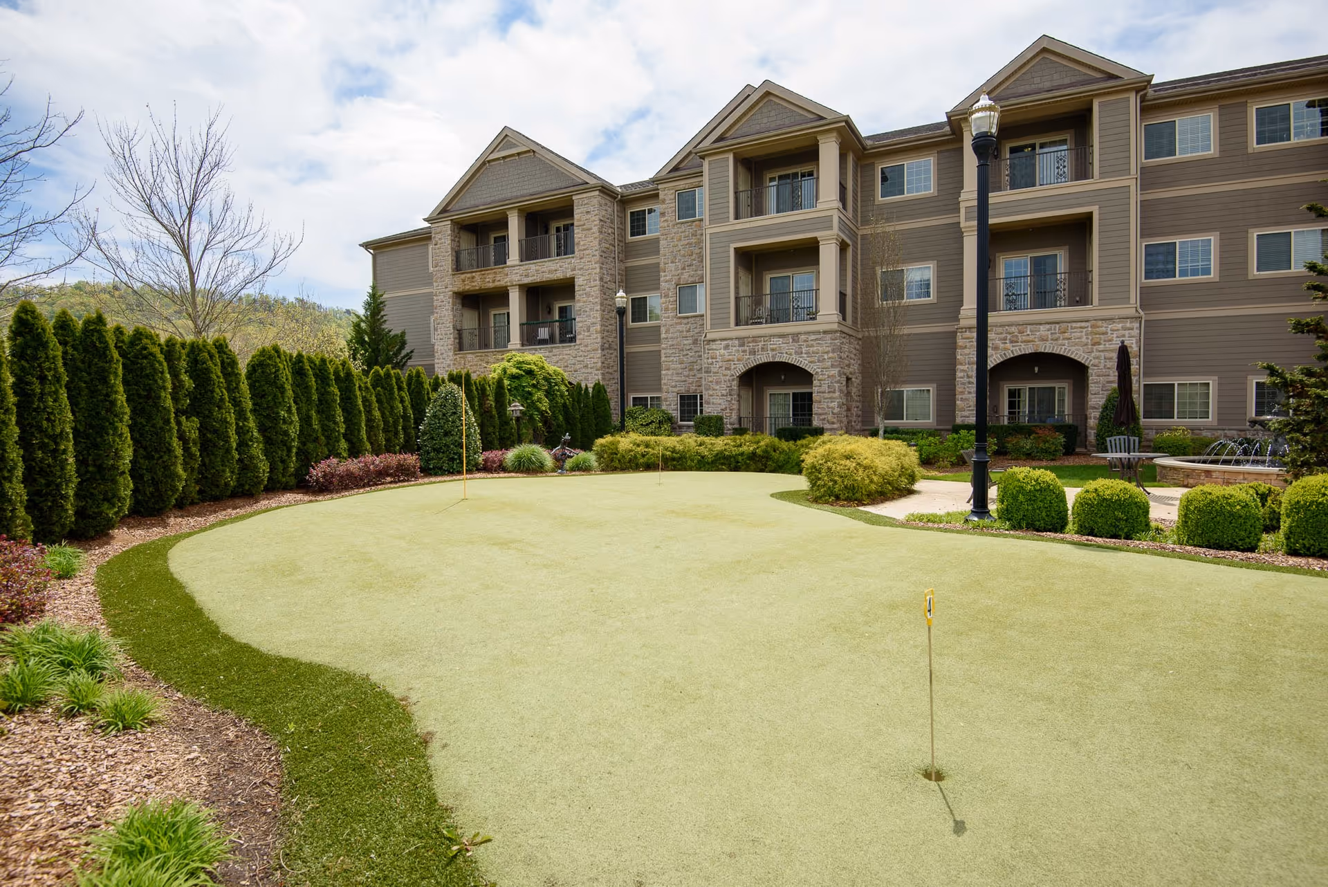 Outdoor putting green surrounded by well-maintained shrubs and trees, with a three-story residential building in the background featuring balconies and stone accents.