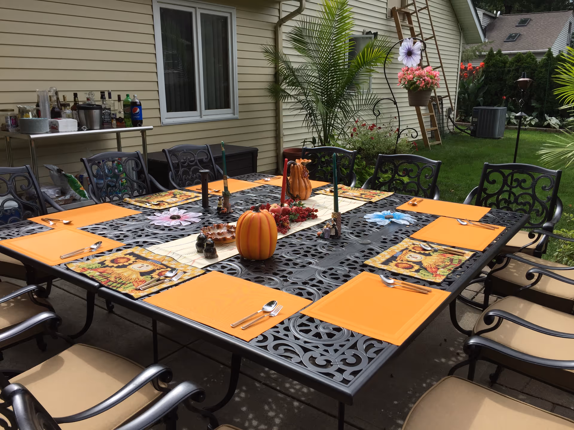 Outdoor patio dining area with a large black metal table set for eight people. The table has orange placemats, sunflower-themed napkins, silverware, and decorative items including a pumpkin centerpiece and candles. The patio is adjacent to a beige house with a window and surrounded by green plants and grass.