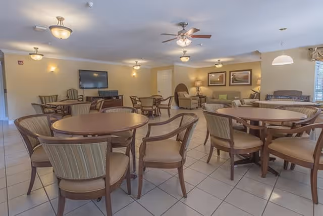A spacious common area with multiple round wooden tables and cushioned chairs arranged on a tiled floor. The room is well-lit with ceiling lights and a ceiling fan. In the background, there is a seating area with armchairs, a lamp, framed artwork on the wall, and a flat-screen TV mounted on the wall.