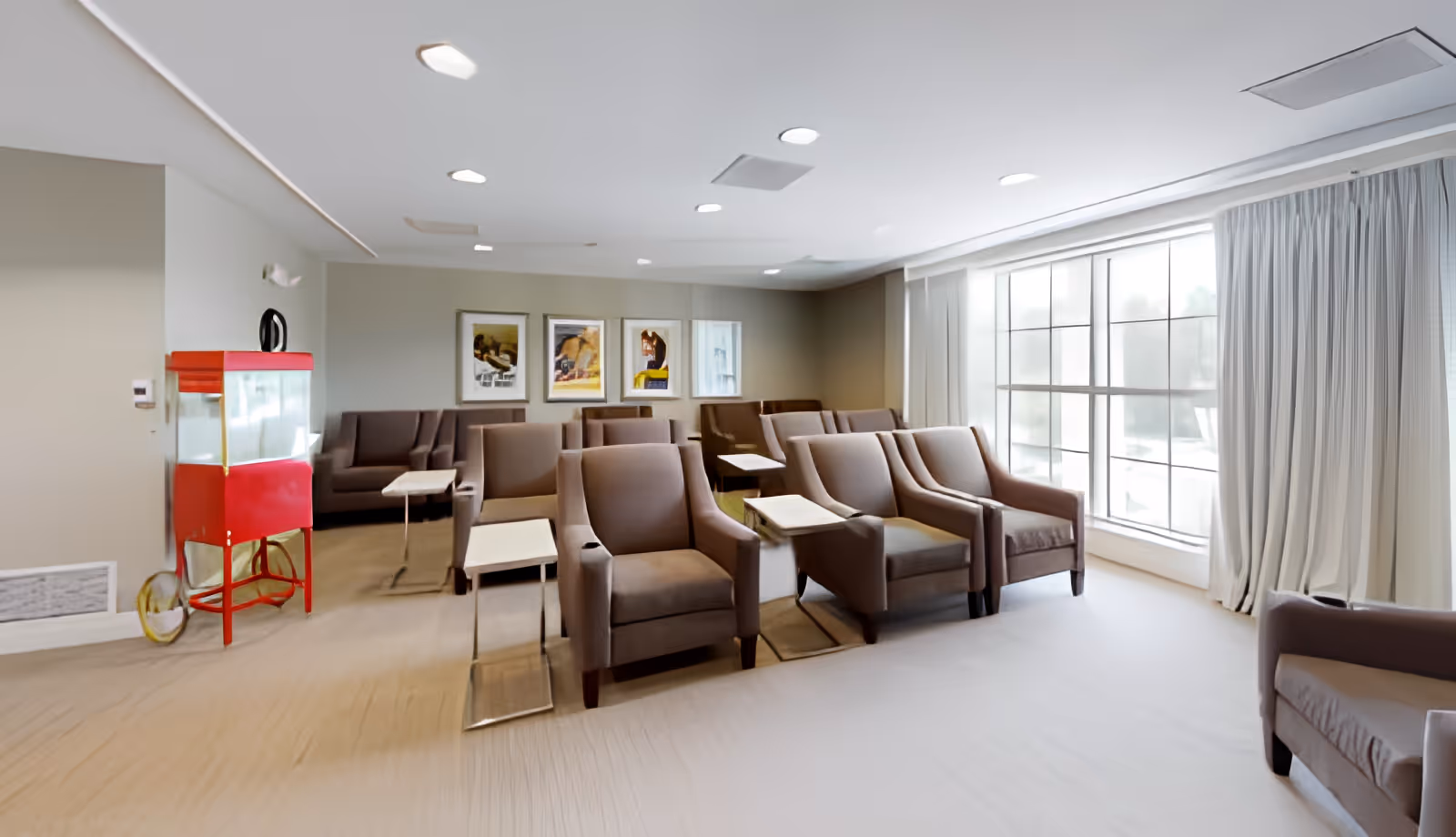 A bright and spacious common area with multiple brown armchairs arranged in rows, each accompanied by small side tables. Large windows with white curtains allow natural light to fill the room. On the left side, there is a red popcorn machine. The walls are decorated with framed artwork.
