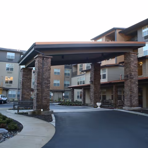 Entrance of a senior living facility with a covered driveway supported by stone pillars. The building has multiple stories with windows and balconies, and there is a curved sidewalk and landscaped area with shrubs and rocks.