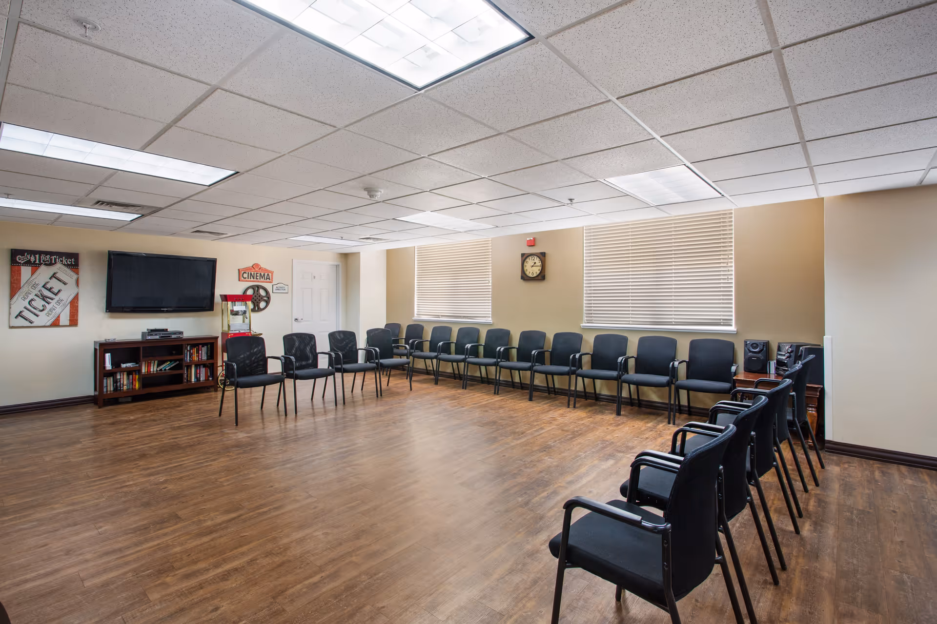 A large room with wooden flooring and beige walls, arranged with black chairs lined up along two walls forming a U shape. The room has a wall-mounted flat-screen TV, a popcorn machine, a bookshelf with books, and movie-themed wall decorations including a ticket and a cinema sign. Two windows with blinds and a clock on the wall are also visible.