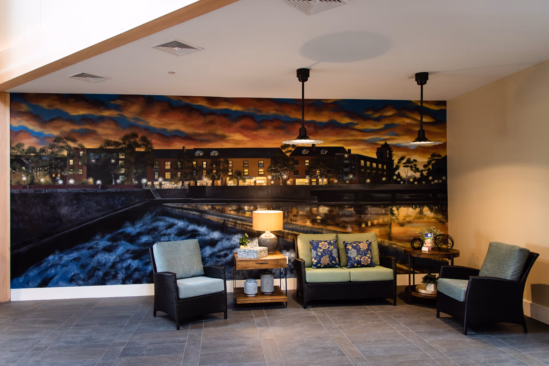 A cozy seating area with two armchairs and a loveseat with green cushions and floral pillows, arranged around two wooden side tables with a lamp and decorative items. Behind the seating area is a large mural depicting a nighttime riverside scene with buildings and trees under a dramatic sky.