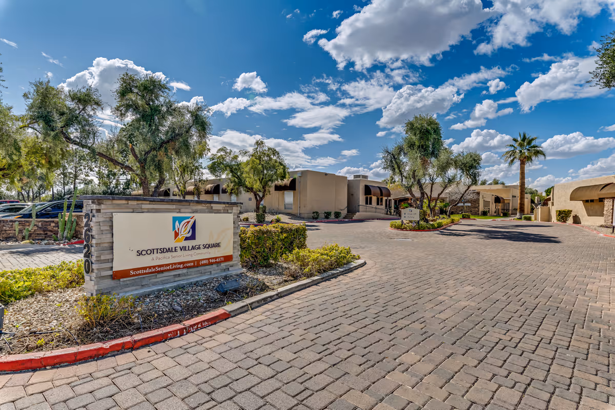 Entrance area of Scottsdale Village Square senior living community with a large sign displaying the community name and contact information. The scene includes a paved driveway, landscaped greenery, trees, and single-story buildings under a partly cloudy blue sky.