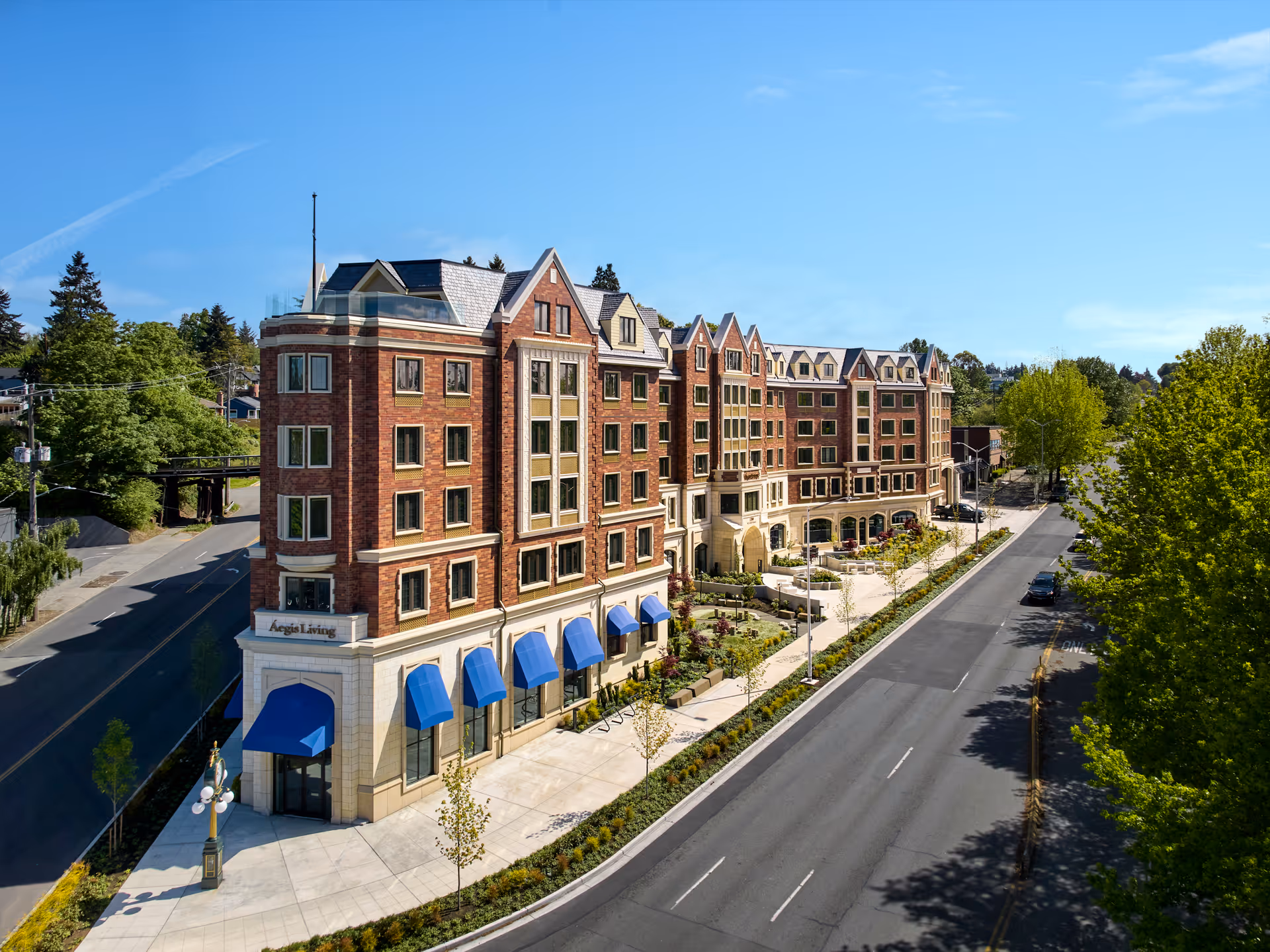 Exterior view of a large multi-story brick building with blue awnings over the windows on the ground floor, labeled Aegis Living. The building is situated along a wide street with trees and greenery surrounding it under a clear blue sky.