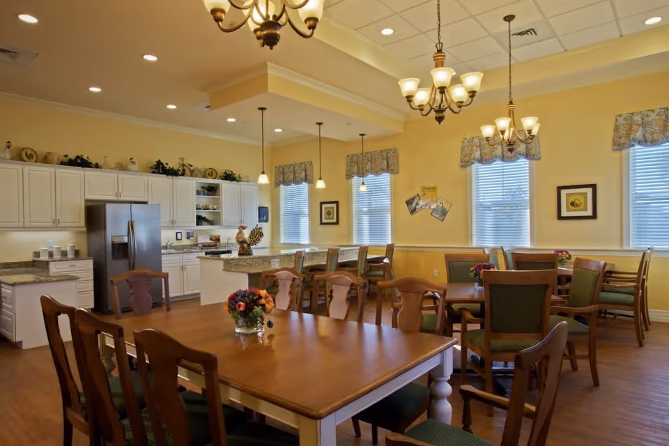 A bright and spacious dining area with wooden tables and chairs, decorated with small flower arrangements. The room features a kitchen area with white cabinets, granite countertops, a stainless steel refrigerator, and pendant lighting. Yellow walls with windows covered by patterned valances allow natural light to fill the space.