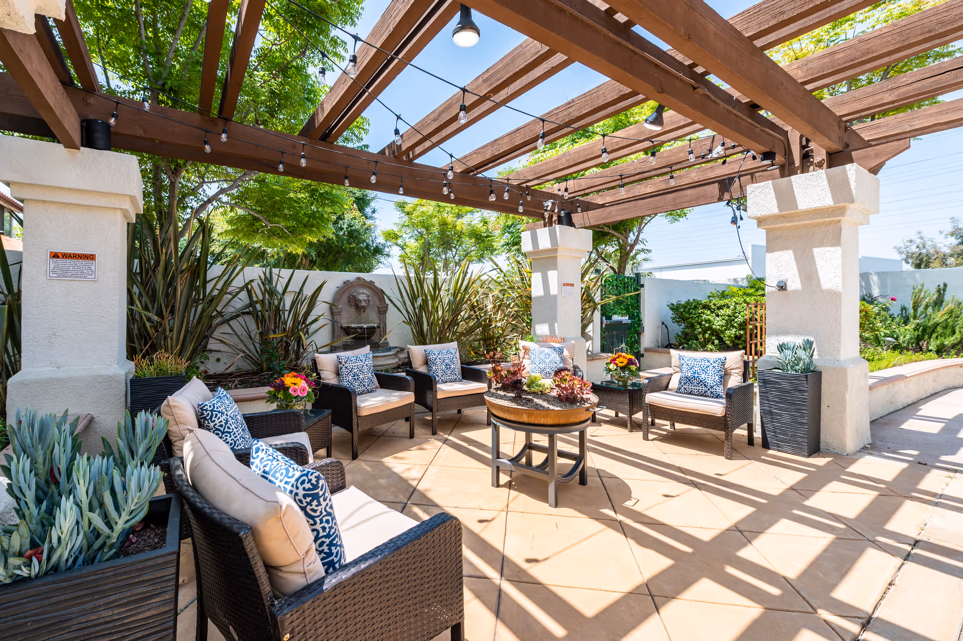 Outdoor seating area with wicker chairs and beige cushions arranged in a circle under a wooden pergola with string lights. The space is surrounded by plants and trees, with a decorative wall fountain in the background and a round table with plants in the center.