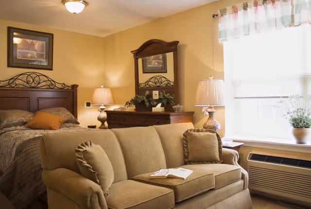 A cozy bedroom with a beige sofa in the foreground, a bed with a decorative headboard and pillows, two bedside tables with lamps, a wooden dresser with a mirror, a framed picture on the wall, and a window with sheer curtains letting in natural light.