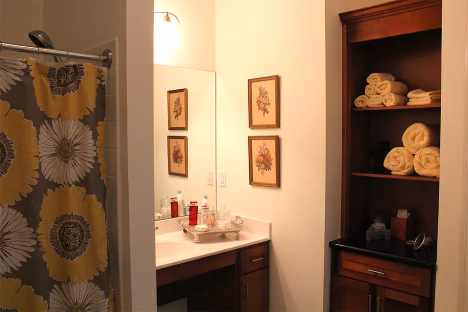 Bathroom with a shower curtain featuring large yellow and white flowers, a vanity with a sink, toiletries on a tray, a large mirror, two framed floral pictures on the wall, and a wooden shelving unit stocked with rolled and folded towels.