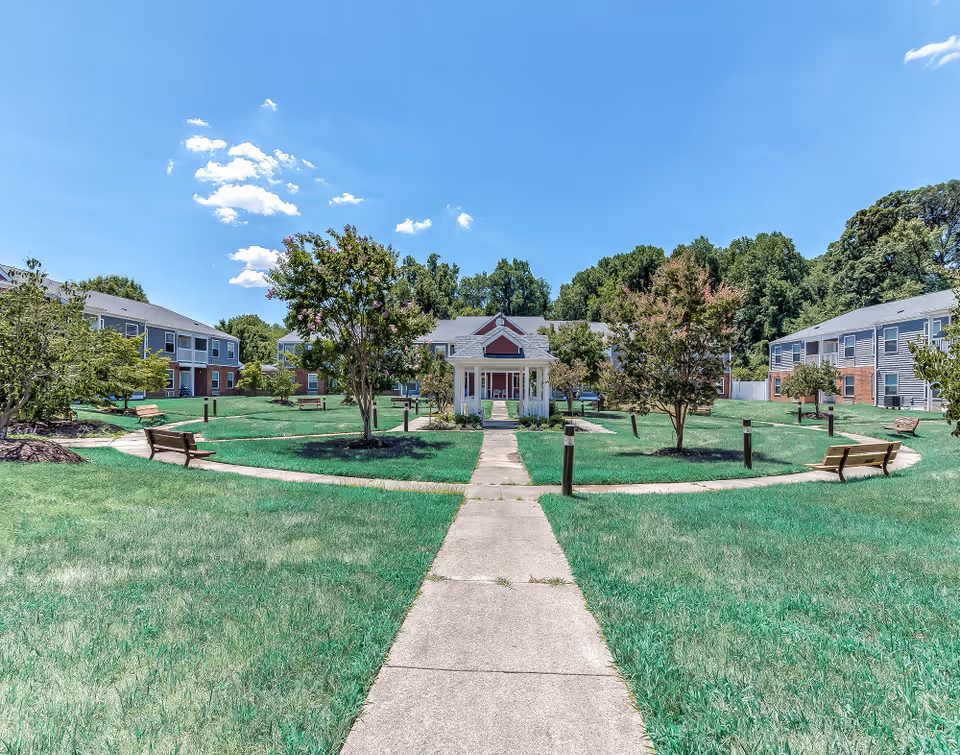 Outdoor courtyard area at Rollingcrest Commons featuring a central gazebo surrounded by green grass, trees, benches, and pathways. Residential buildings with two stories are visible in the background under a clear blue sky with a few clouds.