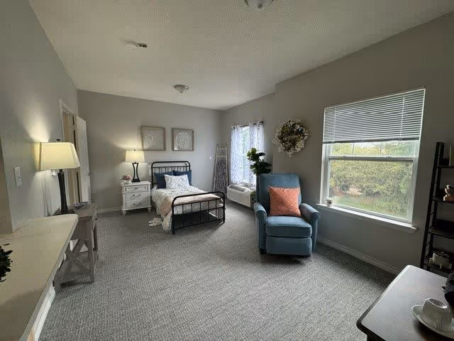 A spacious senior living bedroom with a single metal bed, white bedding, and a nightstand with a lamp. There is a blue armchair with a coral pillow near a large window with blinds and a sheer curtain. The room has light gray walls, carpeted floor, a small table with a lamp, a decorative wreath on the wall, and a shelving unit with decor items.