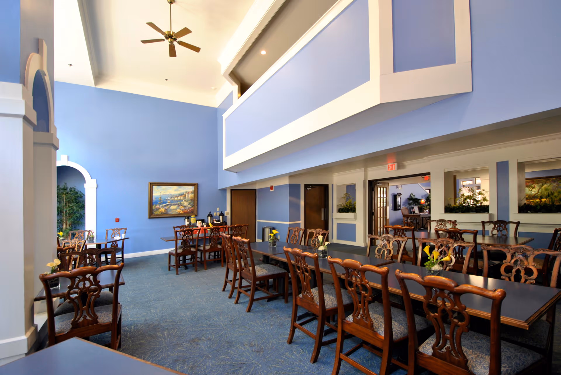 A spacious dining room with blue walls and carpet, featuring multiple wooden tables and chairs arranged neatly. The room has a high ceiling with a ceiling fan, decorative plants, and framed artwork on the walls. There are windows and doorways leading to other areas of the facility.