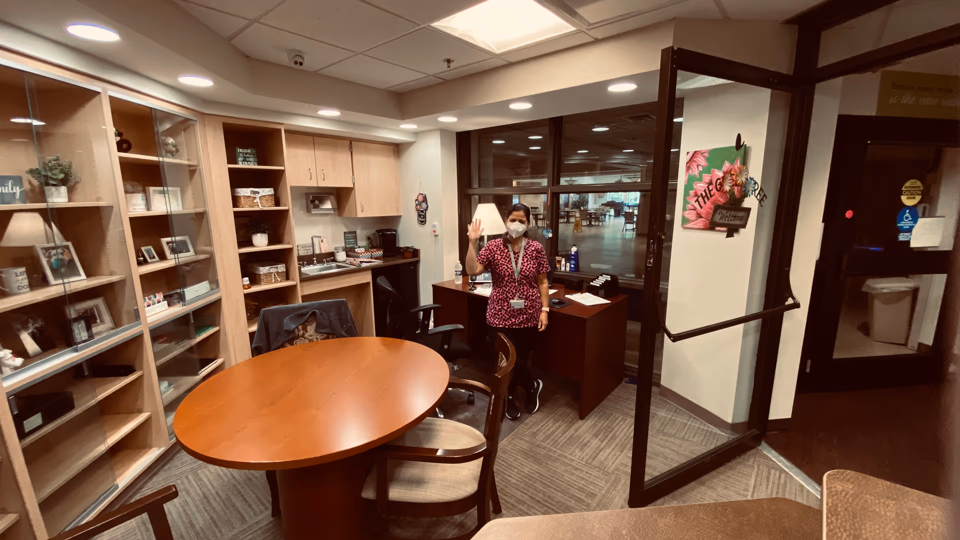 A woman wearing a face mask and a patterned scrub top stands behind a desk in a small office area with wooden shelves, cabinets, and a round wooden table with chairs. The office is well-lit with ceiling lights, and there is a glass door leading to a larger room in the background.