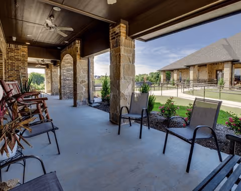 Covered outdoor patio area with multiple chairs and rocking chairs arranged along the walkway. Stone pillars support the roof, and there is a view of a garden with flowers and a building in the background under a clear blue sky.