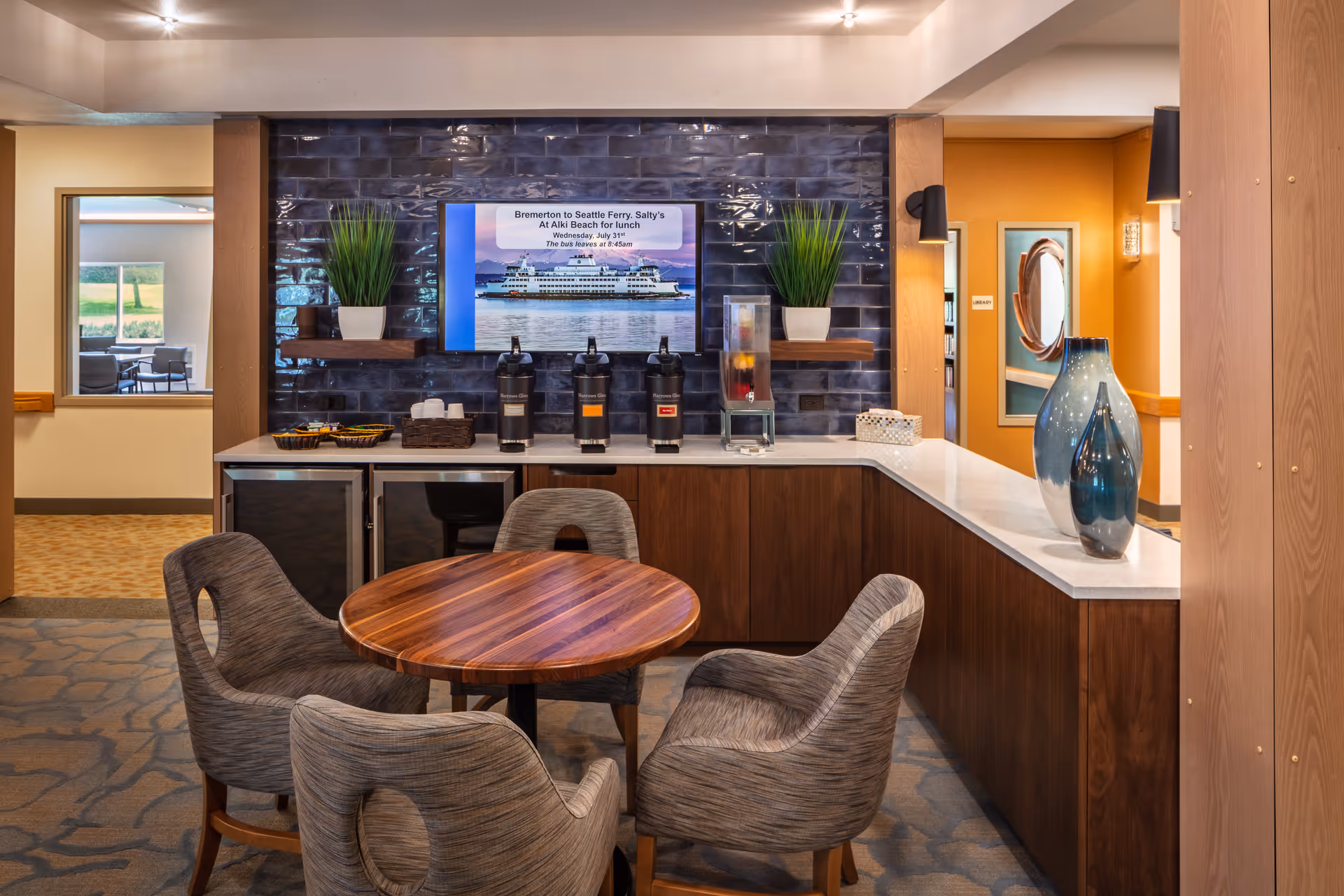 A cozy seating area in a senior living facility featuring a round wooden table surrounded by four cushioned chairs. Behind the table is a countertop with coffee dispensers, a water dispenser, and decorative plants on shelves against a dark tiled wall. A flat-screen TV mounted on the wall displays information about a ferry trip. The room has warm lighting and a modern, inviting atmosphere.