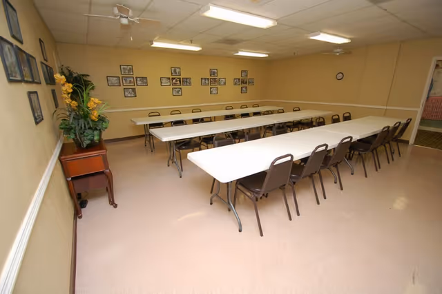 A meeting or activity room with two long white tables arranged in a U-shape surrounded by brown chairs. The walls are beige with a white chair rail, decorated with framed photos and a large plant on a wooden stand. The room has a tiled ceiling with fluorescent lights and a wall clock.
