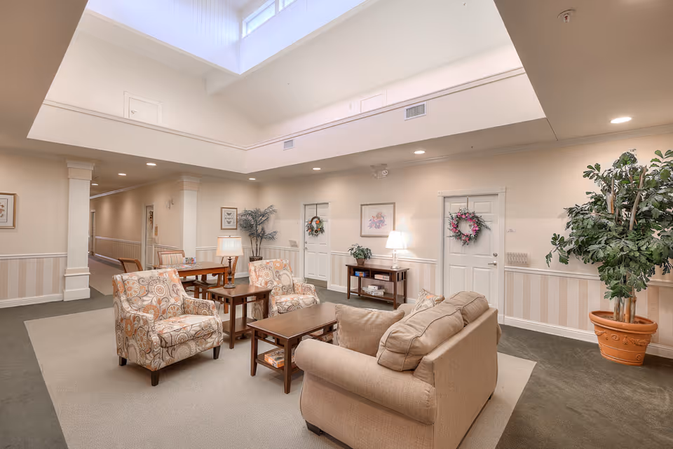 A bright communal seating area with a sofa, patterned armchairs and coffee tables under a skylight in a senior living facility.