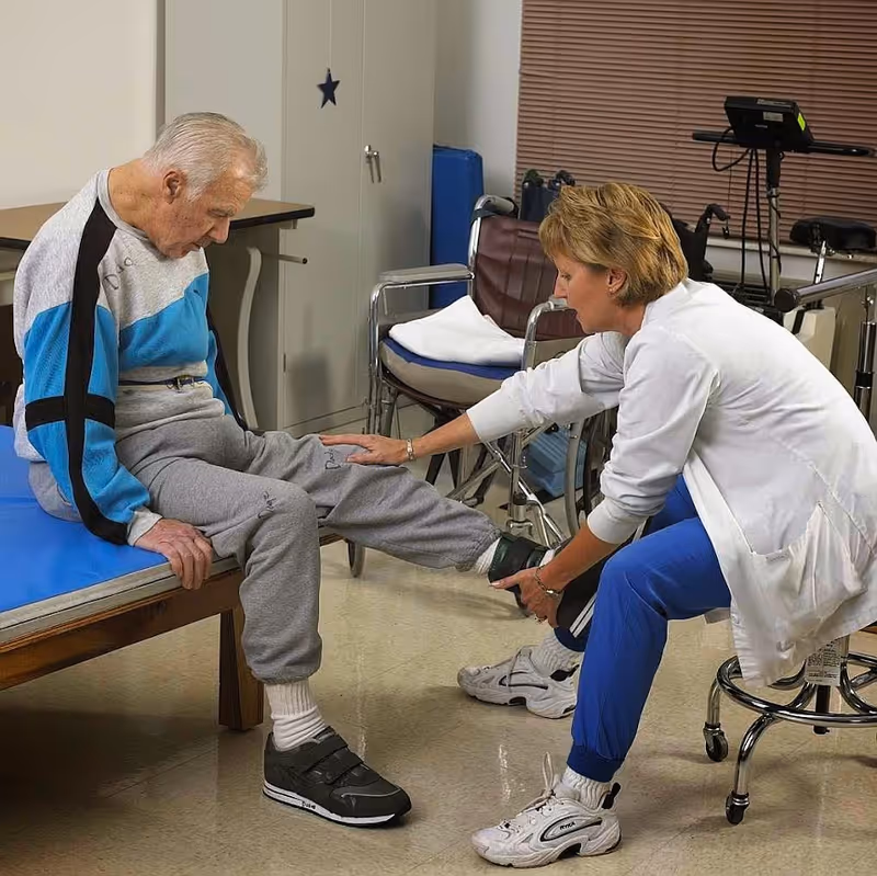A healthcare professional in a white coat and blue pants is assisting an elderly man seated on a medical examination table by holding and examining his leg. The room contains medical equipment, a wheelchair, and a cabinet in the background.